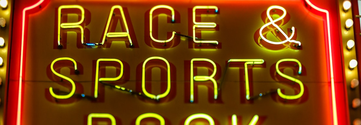 Bright neon lights on a marquee sign display the words 'RACE & SPORTS BOOK'. The sign is surrounded by rows of yellow and white lights against a red background, evoking a vibrant, energetic atmosphere typically associated with casinos or entertainment venues.