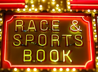 Bright neon lights on a marquee sign display the words 'RACE & SPORTS BOOK'. The sign is surrounded by rows of yellow and white lights against a red background, evoking a vibrant, energetic atmosphere typically associated with casinos or entertainment venues.
