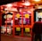 A brightly lit entrance to a casino with neon signs, including one advertising a £500 jackpot. The doors have clear signage indicating entry is restricted to those over 18. A person in a red jacket stands to the right, possibly interacting with a machine or display.