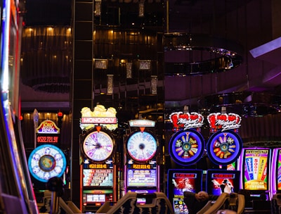 A lively casino scene featuring a row of bright, colorful slot machines with themes like Monopoly, Michael Jackson, and Lord of the Rings. The gaming machines display a variety of flashing lights and digital screens, creating an energetic and exciting atmosphere. Reflections of the lights are visible in glossy surfaces above the machines.