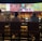 A group of people sit at a bar with their backs to the camera, watching multiple sports games displayed on large screens. The bar counter is lined with various bottles of liquor, and the atmosphere suggests a casual and relaxed social setting.