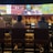A group of people sit at a bar with their backs to the camera, watching multiple sports games displayed on large screens. The bar counter is lined with various bottles of liquor, and the atmosphere suggests a casual and relaxed social setting.