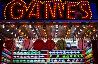 A brightly lit carnival game stall with a large 'GAMES' sign at the top surrounded by red and yellow lights. Various sports balls, including basketballs and soccer balls, are hanging from the ceiling and neatly arranged on shelves below.